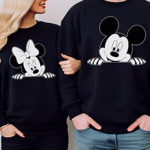May include: Black sweatshirts featuring Mickey and Minnie Mouse designs. The Mickey Mouse sweatshirt has a smiling face with hands peeking over the bottom. The Minnie Mouse sweatshirt has a bow and smiling face. Both designs are in black and white.