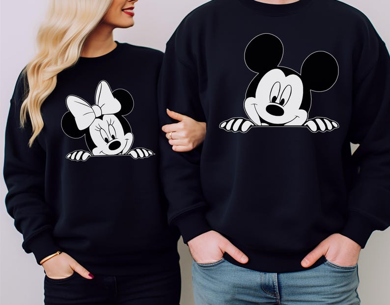 May include: Black sweatshirts featuring Mickey and Minnie Mouse designs. The Mickey Mouse sweatshirt has a smiling face with hands peeking over the bottom. The Minnie Mouse sweatshirt has a bow and smiling face. Both designs are in black and white.