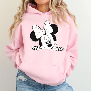 May include: Light pink hooded sweatshirt featuring a black and white Minnie Mouse graphic. The graphic shows Minnie Mouse peeking over the bottom edge of the sweatshirt. The sweatshirt has a front pocket and a drawstring hood. The sleeves are long.