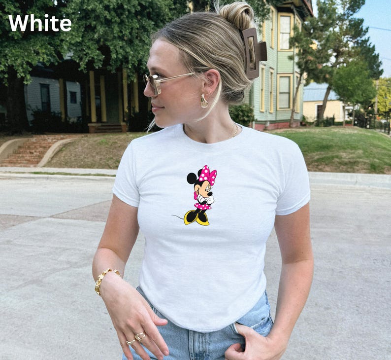 May include: White t-shirt featuring a Minnie Mouse graphic. The cartoon character is depicted in a pink polka-dot bow, dress, and yellow shoes. The shirt has short sleeves and a crew neck. The word "White" is in the top left corner.