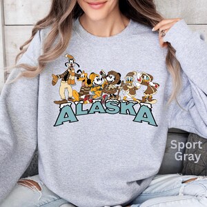 Peut inclure: Sweat-shirt gris sport avec un motif de personnages de dessins anim&eacute;s en v&ecirc;tements d'hiver et le mot "ALASKA" en lettres bleues. Les personnages incluent Dingo, Pluto, Mickey Mouse, Donald Duck, et d'autres.