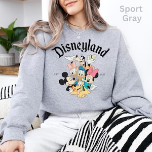 May include: Sport gray sweatshirt featuring the word "Disneyland" in a black script font. Below the word is a graphic of Mickey Mouse, Minnie Mouse, Donald Duck, Daisy Duck, Goofy, and Pluto. The sweatshirt also has the text "EST. 1955".