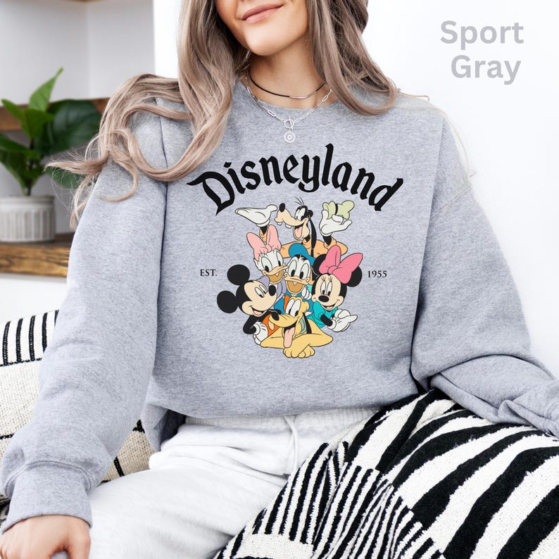 May include: Sport gray sweatshirt featuring the word "Disneyland" in a black script font. Below the word is a graphic of Mickey Mouse, Minnie Mouse, Donald Duck, Daisy Duck, Goofy, and Pluto. The sweatshirt also has the text "EST. 1955".