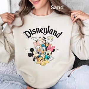 May include: Sand-colored Disneyland sweatshirt featuring a graphic of Mickey Mouse, Minnie Mouse, Donald Duck, Daisy Duck, Goofy, and Pluto. The word "Disneyland" is in a black script font above the characters, with "EST. 1955" below.
