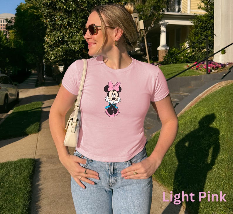 May include: Pale pink t-shirt with a Minnie Mouse design. The top has short sleeves and a round neck, worn with blue jeans. The words "Light Pink" are at the bottom of the image.