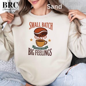 May include: A sand-colored crewneck sweatshirt featuring a graphic print with the text "SMALL BATCH" above a coffee bean and a steaming cup, with the text "ROASTED DAILY" on a green banner and "BIG FEELINGS" below, accented by two small stars.