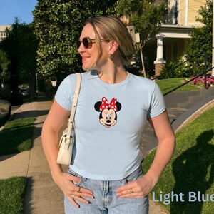 May include: Light blue t-shirt featuring a Minnie Mouse graphic with a red bow and black polka dots. The shirt is paired with blue jeans. The text "Light Blue" is visible in the image.