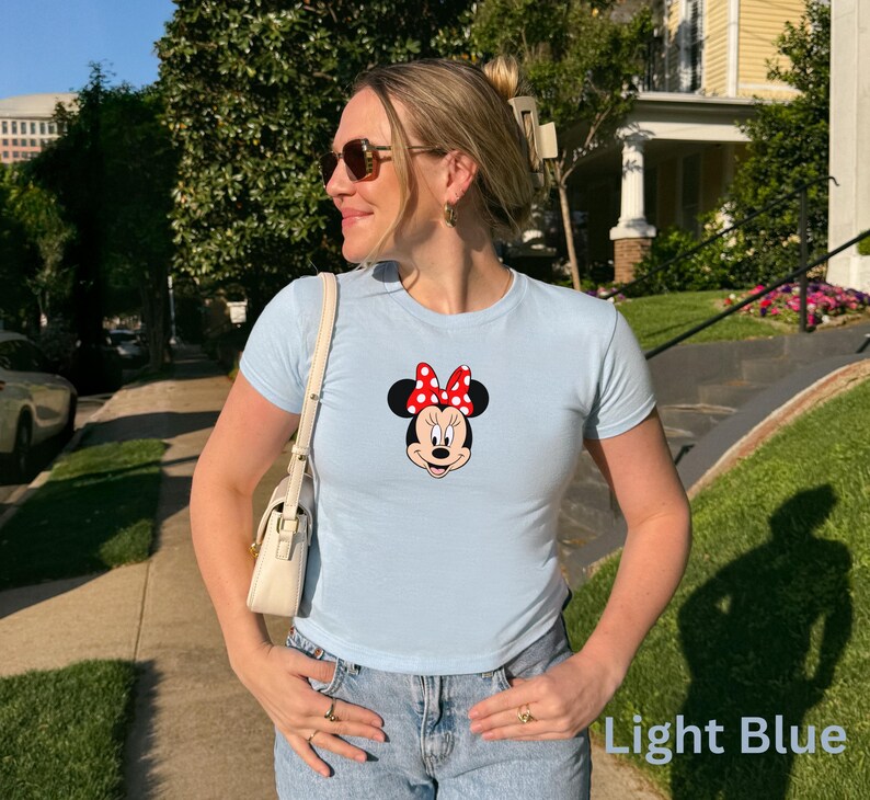 May include: Light blue t-shirt featuring a Minnie Mouse graphic with a red bow and black polka dots. The shirt is paired with blue jeans. The text "Light Blue" is visible in the image.