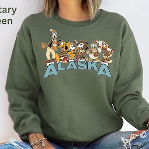 Peut inclure: Sweat-shirt vert militaire avec un motif de personnages Disney, dont Dingo, Mickey Mouse et Donald Duck, et le mot "ALASKA" en lettres bleues. Le sweat-shirt est de style col rond.