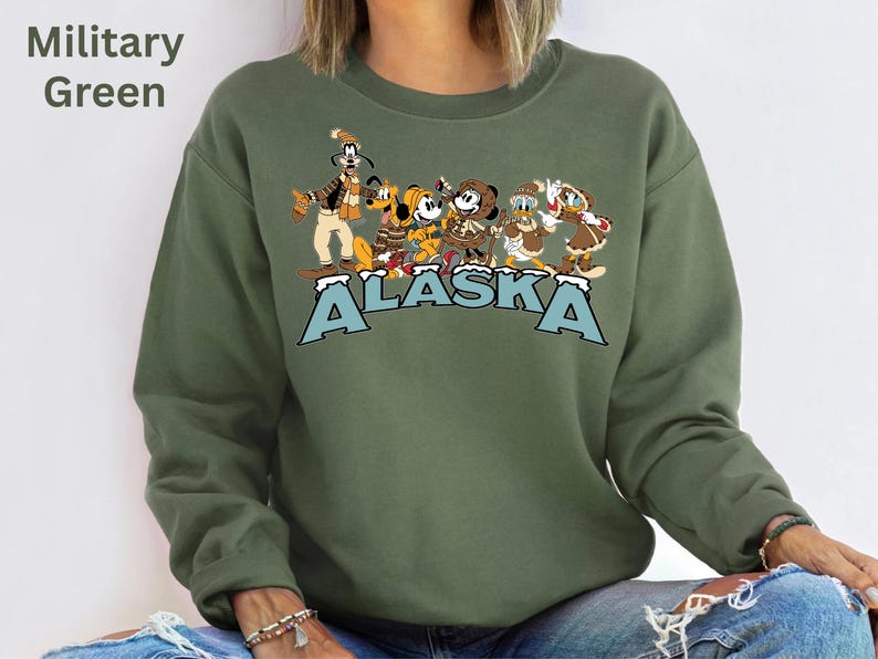 Peut inclure: Sweat-shirt vert militaire avec un motif de personnages Disney, dont Dingo, Mickey Mouse et Donald Duck, et le mot "ALASKA" en lettres bleues. Le sweat-shirt est de style col rond.