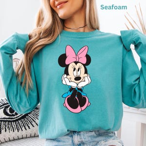 May include: Seafoam green sweatshirt featuring a Minnie Mouse graphic. The cartoon character is depicted with a pink bow, blue dress, and white gloves. The word "Seafoam" is printed at the top of the image.