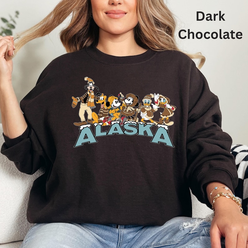 Peut inclure: Sweat-shirt chocolat fonc&eacute; avec un motif de personnages de dessins anim&eacute;s en v&ecirc;tements d'hiver. Le mot "ALASKA" est imprim&eacute; en bleu et blanc. Le sweat-shirt est de couleur marron fonc&eacute;.