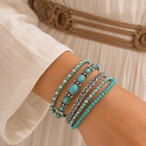 Könnte beinhalten: Ein Set aus vier Armbändern mit türkisfarbenen Perlen, ovalen türkisfarbenen Steinen in silberfarbenen Fassungen und einem silberfarbenen Metallband mit einem komplizierten Ausschnittmuster, die zusammen am Handgelenk getragen werden.