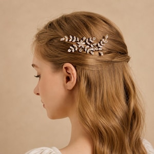 May include: A gold-toned hair comb featuring a delicate branch and leaf design. The accessory is embellished with numerous small, clear, sparkling crystals arranged along the metallic stems, creating a botanical-inspired hair ornament.