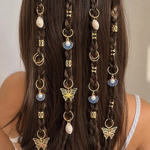 Puede incluir: Cuatro trenzas de cabello castaño decoradas con anillos, puños y dijes de metal dorado. Los dijes incluyen conchas marinas blancas, conchas de abanico de esmalte azul y blanco con perlas, y dijes de mariposa dorados en anillos.