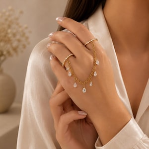 May include: A gold-toned hand chain jewelry piece featuring two thin bands connected by a delicate chain. The chain is adorned with multiple small, clear, teardrop-shaped stones that hang down, creating a draped effect across the back of the hand.