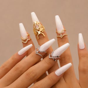 May include: A collection of five rings worn on fingers with long white nails. The set includes gold-toned rings with filigree and chevron designs, and silver-toned chevron rings, all featuring small clear stones set along the bands.