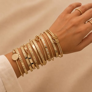 Puede incluir: Una colección de pulseras doradas en la muñeca, con diversos diseños que incluyen bandas lisas, superficies texturizadas, metal trenzado y pequeñas piedras incrustadas. Dos anillos dorados sencillos se llevan en los dedos de la mano.