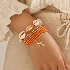 May include: A multi-strand bracelet set featuring one strand of white cowrie shells on an orange cord and several strands of orange, yellow, and gold-toned seed beads. A gold-toned starfish charm hangs from the beaded section of the bracelet.