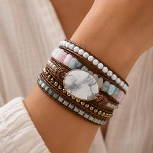 Peut inclure: Un bracelet multi-rangs doté d'une grande pierre centrale ovale au motif marbré blanc et gris. Le design intègre des rangées de perles blanches, des pierres rose pâle et bleu clair, des sphères dorées et des perles grises à facettes.