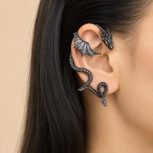 May include: A dark metallic ear cuff featuring a detailed dragon design that wraps around the ear. The piece includes a dragon head at the top, a textured wing extending across the upper ear cartilage, and a long, scaled tail curving downward.