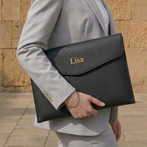 May include: Black leather portfolio with a flap closure, embossed with the name "Lisa" in gold lettering. The portfolio is held by a person wearing a gray suit. A gold bracelet is visible on the wrist.