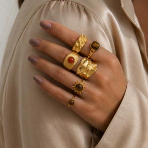 Puede incluir: Una colección de seis anillos dorados usados en los dedos. El conjunto incluye anillos con texturas orgánicas irregulares, un anillo con una piedra naranja redonda, un anillo con una piedra marrón cuadrada y dos anillos con diseños de rayos de sol con piedras marrones.