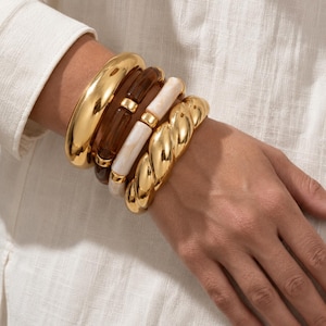 May include: A stack of four bracelets featuring a smooth polished gold bangle, a dark brown resin link bracelet with gold accents, a cream-colored resin link bracelet with gold accents, and a thick twisted gold metal cuff bracelet.