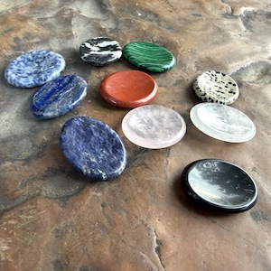 May include: A collection of oval-shaped polished stones in various colors and patterns. The stones include blue, green, red, black, and white, with unique textures and veining. The arrangement is on a textured, brown surface.