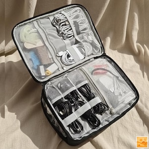 Peut inclure: Organisateur de voyage noir ouvert avec plusieurs compartiments. Il contient divers accessoires électroniques, notamment des câbles blancs et noirs, des écouteurs et de petits gadgets. L'intérieur comprend des poches en filet et des sangles élastiques.