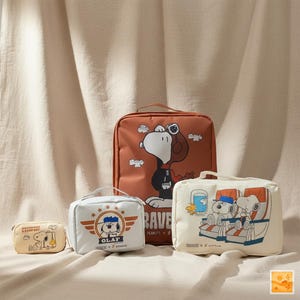 May include: Four travel bags featuring Snoopy and other Peanuts characters. The bags are in various sizes and colors, including tan, white, and orange. One bag has Snoopy in a pilot outfit, and another has Snoopy and Woodstock on an airplane.