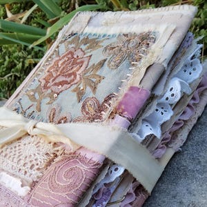 May include: A handmade fabric journal featuring a cover with floral embroidery, sequins, and layered lace. The book is bound with a cream-coloured ribbon tie and includes various textured fabric pages, eyelet lace trim, and pink velvet accents.