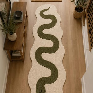 May include: An elongated, cream-coloured area rug with an irregular, wavy edge. A long, winding dark olive green snake graphic runs down the centre of the rug. The snake features a rounded head with a small black eye and a black forked tongue detail.
