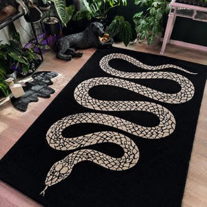 May include: A rectangular area rug featuring a large, winding snake design in a light beige color against a solid black background. The snake pattern displays a detailed, textured scale motif throughout its entire length, extending from the head.