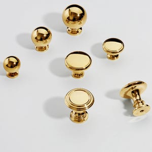 Op de afbeelding: Een verzameling goudkleurige kastknoppen in verschillende vormen en maten. De knoppen omvatten bolvormige en platte ontwerpen, allemaal met een glanzende, reflecterende afwerking. De hardware is gerangschikt op een wit oppervlak, wat hun gepolijste uiterlijk benadrukt.