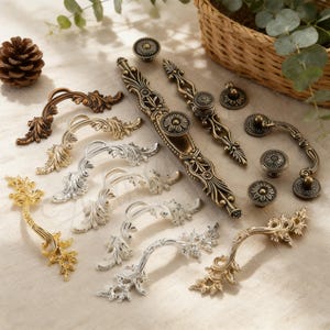 May include: An assortment of decorative cabinet hardware in various metallic finishes, including gold, silver, and bronze. The hardware features ornate, floral-inspired designs, including handles and knobs. A pinecone and a woven basket are in the background.