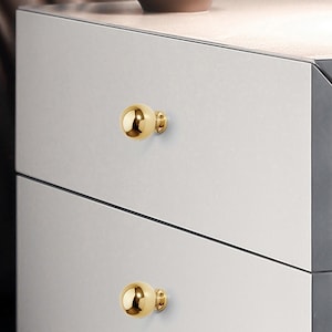 Op de afbeelding: Close-up van een moderne dressoir met twee lades. Elke lade heeft een glanzende, goudkleurige, bolvormige knop. De knoppen zijn bevestigd aan de lichtgrijze ladefronten. De dressoir staat tegen een neutrale achtergrond.
