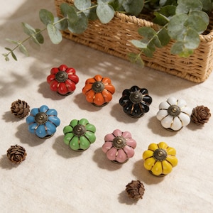 May include: A collection of colorful ceramic cabinet knobs in various shades, including red, orange, blue, green, pink, and yellow. Each knob has a flower-like design with a bronze-colored center. The knobs are arranged on a neutral surface.