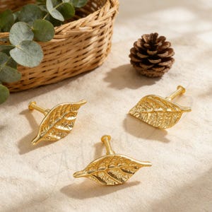 Op de afbeelding: Drie goudkleurige, bladvormige kastknoppen met gedetailleerde nerven. De knoppen zijn gerangschikt op een neutraal oppervlak, met een rieten mand, een dennenappel en groen in de achtergrond. Deze decoratieve knoppen voegen een natuurlijke toets toe.