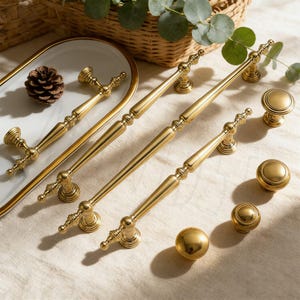 Op de afbeelding: Een verzameling goudkleurige kastbeslagen, waaronder lange stangtrekkers, ronde knoppen en een bolvormige knop. De hardware is gerangschikt op een lichtgekleurd oppervlak, met een decoratieve tray en een dennenappel op de achtergrond. De hardware is gemaakt van metaal.