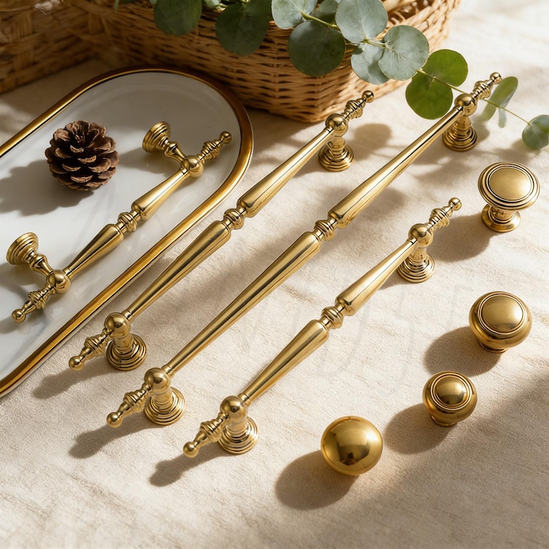 Op de afbeelding: Een verzameling goudkleurige kastbeslagen, waaronder lange stangtrekkers, ronde knoppen en een bolvormige knop. De hardware is gerangschikt op een lichtgekleurd oppervlak, met een decoratieve tray en een dennenappel op de achtergrond. De hardware is gemaakt van metaal.