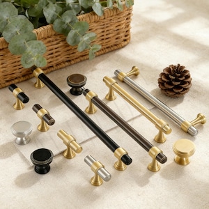 Puede incluir: Una colección de tiradores y pomos para armarios en varios acabados. Los herrajes presentan detalles dorados, plateados y negros. Los tiradores miden unos 15 cm de largo. Una piña y una cesta de mimbre con vegetación en el fondo.