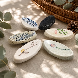 May include: A collection of ceramic chopstick rests in various designs. Each rest is oval-shaped and features hand-painted details, including wave patterns, floral motifs, and abstract designs. The color palette includes white, blue, black, and earth tones.