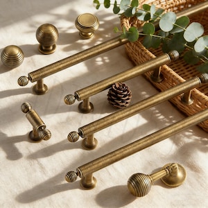 Puede incluir: Un surtido de herrajes de gabinete de latón antiguo, que incluyen perillas y tiradores. Las perillas tienen un diseño redondo y acanalado, mientras que los tiradores tienen forma de barra con extremos decorativos. El hardware está dispuesto sobre una superficie de color neutro.