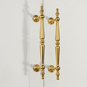 Op de afbeelding: Twee sierlijke, goudkleurige deurklinken. Elke klink heeft een lange, slanke behuizing met decoratieve details en een afgeronde basis. De klinken zijn gemonteerd op een wit oppervlak, wat een contrast cre&euml;ert met de glanzende gouden afwerking.