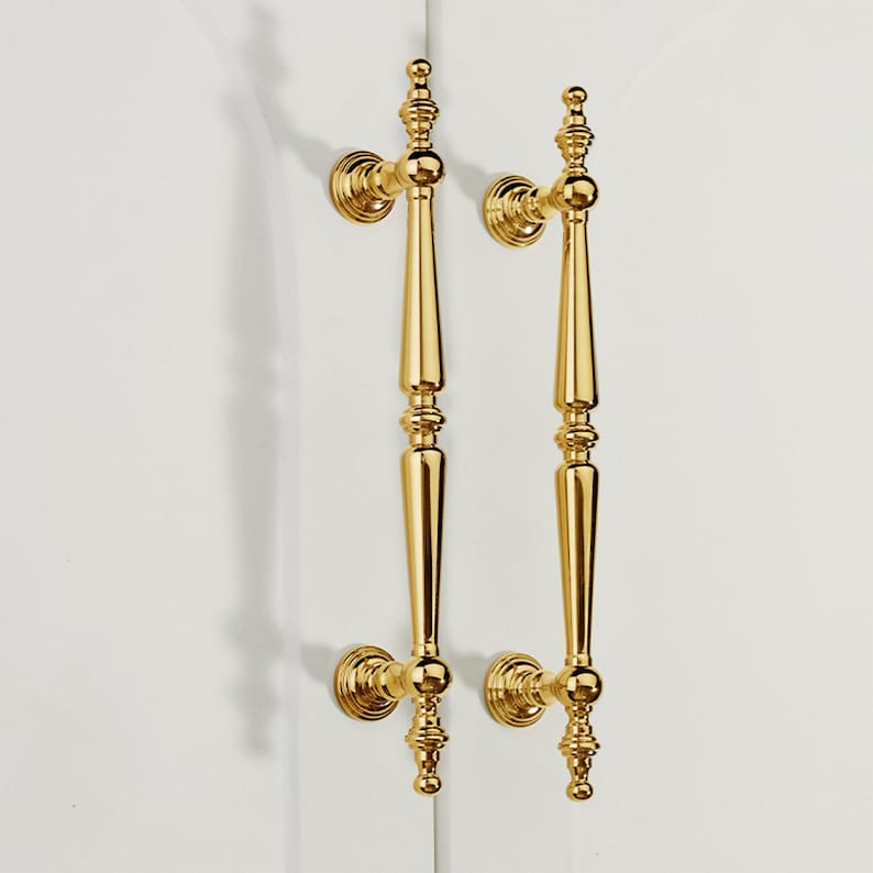 Op de afbeelding: Twee sierlijke, goudkleurige deurklinken. Elke klink heeft een lange, slanke behuizing met decoratieve details en een afgeronde basis. De klinken zijn gemonteerd op een wit oppervlak, wat een contrast cre&euml;ert met de glanzende gouden afwerking.