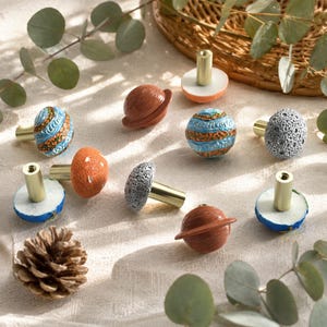 May include: A collection of decorative cabinet knobs in various designs, including spheres, planets, and mushroom shapes. The knobs feature gold-colored stems and are painted in shades of blue, orange, brown, and gray. The knobs are arranged on a cream-colored surface.