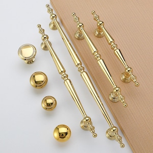 Op de afbeelding: Een verzameling goudkleurige kastbeslagen, waaronder knoppen en handgrepen. De handgrepen zijn lang en slank, met decoratieve details. De knoppen zijn rond en hebben een eenvoudig, elegant ontwerp. De hardware is gerangschikt op een lichtgekleurd houten oppervlak.