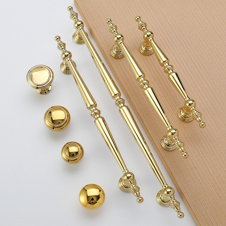 Op de afbeelding: Een verzameling goudkleurige kastbeslagen, waaronder knoppen en handgrepen. De handgrepen zijn lang en slank, met decoratieve details. De knoppen zijn rond en hebben een eenvoudig, elegant ontwerp. De hardware is gerangschikt op een lichtgekleurd houten oppervlak.