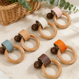 May include: Wooden teething rings with leather accents in various colors, including blue, brown, orange, pink, and white. Each ring is paired with a small pinecone. The rings are arranged on a cream-colored surface.